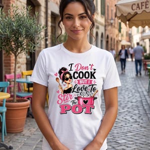 Puede incluir: Una camiseta blanca de manga corta con un estampado gráfico de una persona con diadema rosa y gafas de sol sosteniendo una cuchara de madera, junto a una olla rosa y el texto "I Don't COOK BUT I Love To Stir The POT".