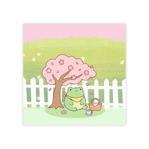 Op de afbeelding: Een digitale illustratie van een groene kikker met een gele halsdoek, zittend onder een roze kersenbloesemboom. Naast de kikker staan een kleine bruine picknickmand en een theekopje. Op de achtergrond staat een wit houten hek.