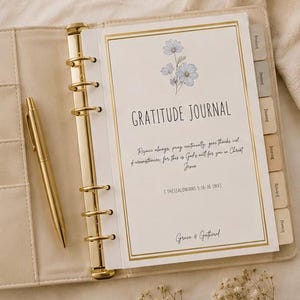 May include: A cream-colored ring-bound gratitude journal with a gold metal spine and a matching gold pen. The cover features a blue floral illustration, the text "GRATITUDE JOURNAL," a Bible verse from 1 Thessalonians 5:16-18, and "Grace & Gathered."