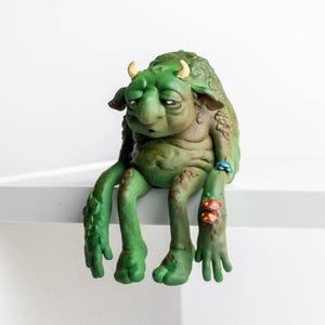 May include: A green, textured creature figurine with small yellow horns, drooping eyes, and a large nose. The figure has long arms and legs, with small colourful mushrooms and blue floral details attached to its limbs, sitting in a slumped posture.