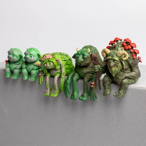Puede incluir: Un conjunto de cinco figuras de criaturas verdes sentadas en fila. Las figuras presentan texturas variadas, incluyendo piel verde musgo, peque&ntilde;os cuernos, coronas de flores, un bast&oacute;n de madera y racimos de setas rojas en sus espaldas.