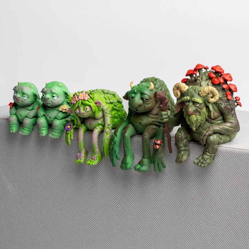Puede incluir: Un conjunto de cinco figuras de criaturas verdes sentadas en fila. Las figuras presentan texturas variadas, incluyendo piel verde musgo, peque&ntilde;os cuernos, coronas de flores, un bast&oacute;n de madera y racimos de setas rojas en sus espaldas.