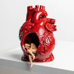 May include: A glossy red anatomical heart sculpture featuring a hollowed-out center. Inside the cavity, a small, beige-toned figurine of a person is curled in a fetal position, with one leg extending outward from the opening of the heart structure.