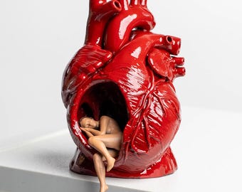 Puede incluir: Una escultura de corazón anatómico de color rojo brillante con un centro hueco. Dentro de la cavidad, una pequeña figura de color beige representa a una persona acurrucada en posición fetal, con una pierna extendida hacia fuera de la abertura.