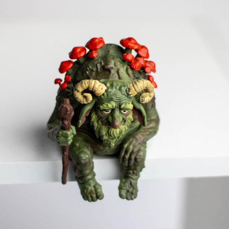 Puede incluir: Una figura de criatura verde y texturizada con cuernos beige curvados, barba y un bast&oacute;n marr&oacute;n. La figura tiene setas de color rojo brillante creciendo en su espalda y cabeza, y est&aacute; representada en posici&oacute;n sentada con las piernas colgando.