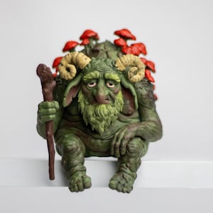 Puede incluir: Una figura de criatura verde y texturizada con cuernos de carnero, barba y orejas grandes, sentada mientras sostiene un bast&oacute;n de madera marr&oacute;n. La figura tiene un grupo de hongos rojos brillantes creciendo en su espalda, con pliegues cut&aacute;neos.