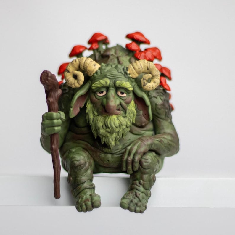 Puede incluir: Una figura de criatura verde y texturizada con cuernos de carnero, barba y orejas grandes, sentada mientras sostiene un bast&oacute;n de madera marr&oacute;n. La figura tiene un grupo de hongos rojos brillantes creciendo en su espalda, con pliegues cut&aacute;neos.