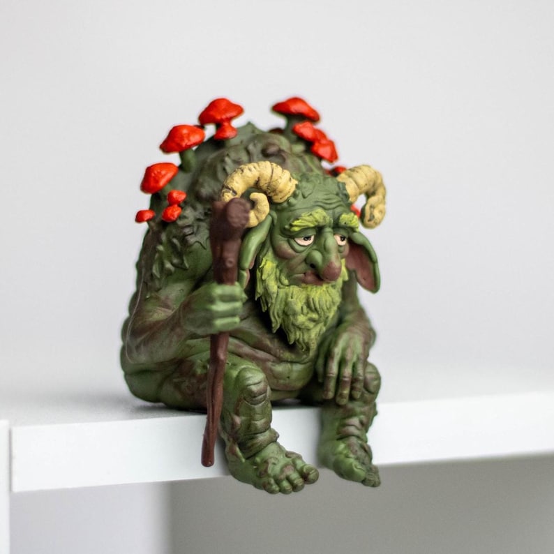 Puede incluir: Una figura de criatura verde y texturizada con cuernos beige curvados, una barba larga y un bast&oacute;n marr&oacute;n. La figura tiene setas de color rojo brillante creciendo en su espalda y est&aacute; sentada encorvada, con pliegues de piel y rasgos detallados.