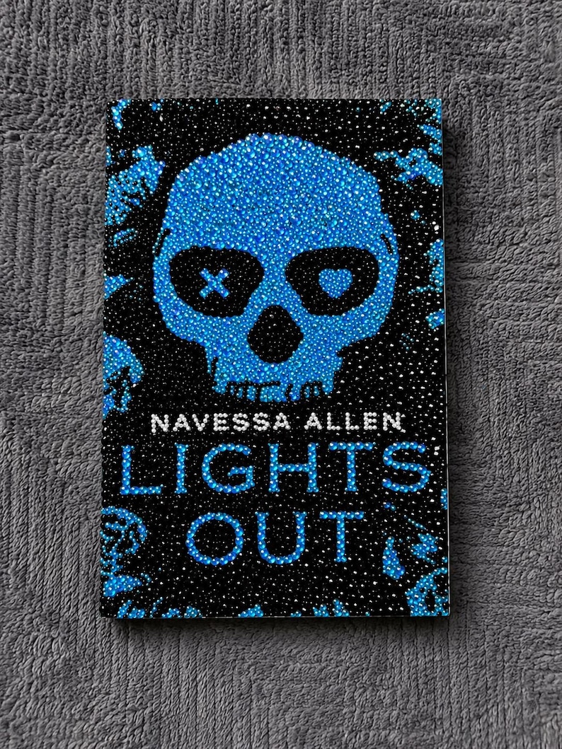 May include: A rectangular book cover titled Lights Out by Navessa Allen, featuring a central blue skull design composed of small rhinestones. The skull has an X and a heart in the eye sockets, set against a black background with blue accents.