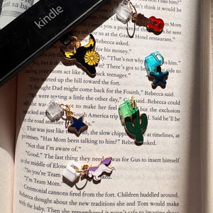 May include: A black Kindle e-reader partially covering a page of text, with five metal charms attached to clear plastic clips. The charms include a red guitar, a black bull head with a yellow flower, a blue star, a green cactus, and a white unicorn.