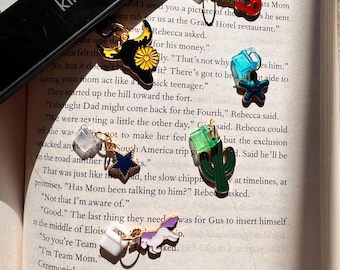 Puede incluir: Un lector electrónico Kindle negro que cubre parcialmente una página de texto, con cinco dijes de metal unidos a clips de plástico transparente. Los dijes incluyen una guitarra roja, una cabeza de toro negra, una estrella azul, un cactus verde.