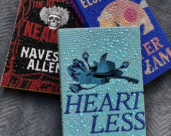 Puede incluir: Tres libros de tapa dura decorados con pedrería. El libro de la izquierda es negro y rojo con un gráfico de calavera. El libro superior es azul con texto dorado que dice ELSIE SILVER y FEVER. El libro frontal es azul claro con un gráfico de guitarra y sombrero y el texto HEARTLESS ELSIE SILVER.