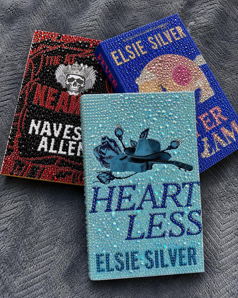 May include: Three hardcover books decorated with rhinestones. The left book is black and red with a skull graphic. The top book is blue with gold text reading ELSIE SILVER and FEVER. The front book is light blue with a guitar and hat graphic and text reading HEARTLESS ELSIE SILVER.