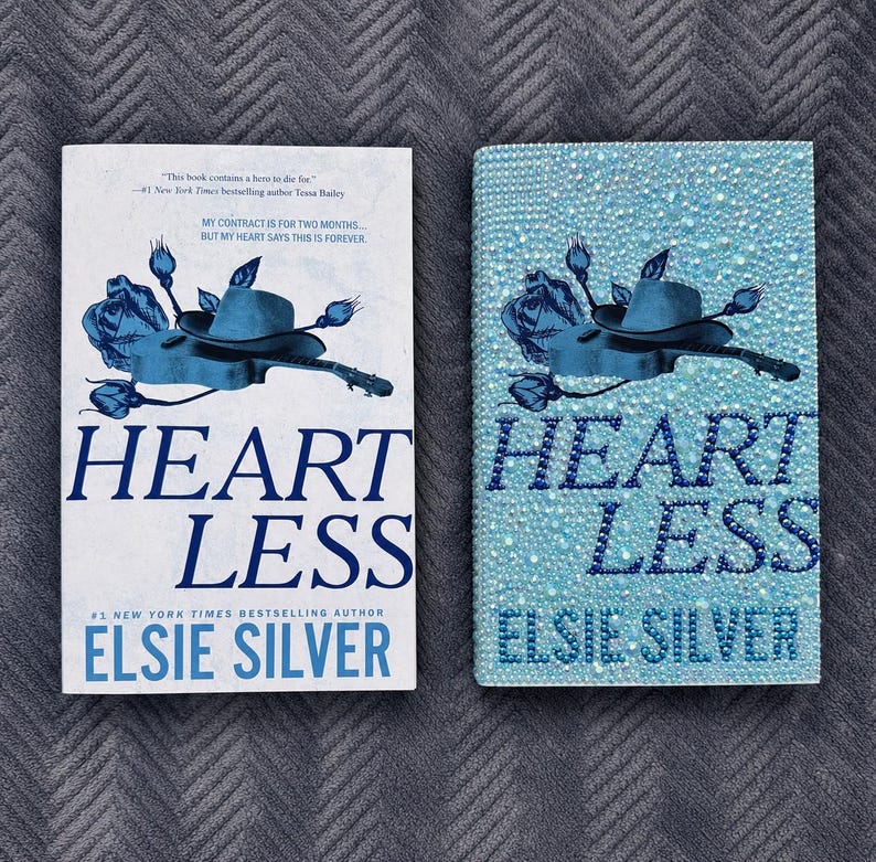 May include: Two copies of the book Heartless by Elsie Silver. The left copy has a white cover with a blue illustration of a hat, guitar, and roses. The right copy is covered in light blue rhinestones, featuring the same blue graphic design.