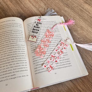 May include: Three rectangular clear acrylic bookmarks with tassels. One features the text "I Like My Books Extra Spicy" with a book charm. Another has a pink bow pattern, and the third displays a grid of red hearts and pink flowers with red bows.