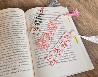 Puede incluir: Tres marcapáginas rectangulares de acrílico transparente con borlas. Uno tiene el texto "I Like My Books Extra Spicy" con un dije de libro. Otro presenta un patrón de lazos rosas y el tercero muestra una cuadrícula de corazones rojos y flores rosas.