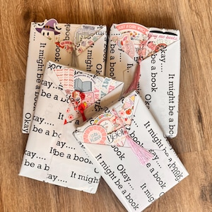 May include: Four rectangular packages wrapped in white paper printed with the repeating black text "Okay... It might be a book". Each package is decorated with colorful stickers, including a ghost in a hat, and features a hanging tassel.