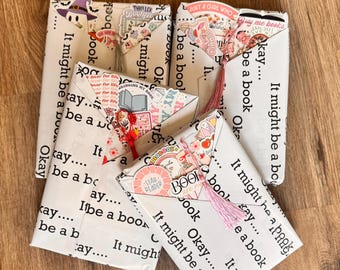 Puede incluir: Cuatro paquetes rectangulares envueltos en papel blanco impreso con el texto negro repetitivo "Okay... It might be a book". Cada paquete está decorado con pegatinas coloridas, incluido un fantasma con sombrero, y tiene una borla colgante.