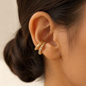 May include: Two gold-toned ear cuffs worn on the cartilage. One cuff features a smooth, polished metal surface, while the other is set with a row of small, clear, sparkling stones. Both pieces are circular and fit snugly around the ear edge.