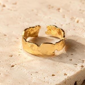 May include: A gold-toned ring with an open band design, featuring a textured, irregular, and hammered surface finish. The metal band has a reflective, polished appearance with uneven edges that create a distinct, organic, and non-uniform shape.