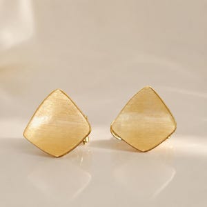 May include: A pair of gold-toned stud earrings featuring a brushed metal finish. Each earring has a distinct four-sided, kite-like geometric shape with soft, rounded corners and a flat, textured surface that reflects light across the metal.