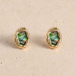 May include: A pair of oval stud earrings featuring a textured gold-tone metal frame surrounding a central inlay of iridescent abalone shell with swirling green, purple, and blue tones. The earrings have a polished finish and a rounded organic shape.