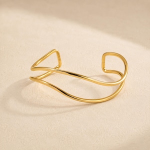 May include: A gold-toned cuff bracelet featuring a double-layered design with smooth, curved lines. The open-ended structure consists of two parallel bands that gently wave and intersect, creating a polished, reflective metallic surface throughout.