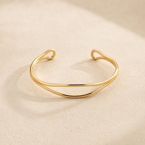 May include: A gold-toned cuff bracelet featuring a double-layered design with two thin, smooth bands that converge at the ends. The open-style bracelet has rounded, looped terminals at each side, creating a continuous, polished metallic appearance.