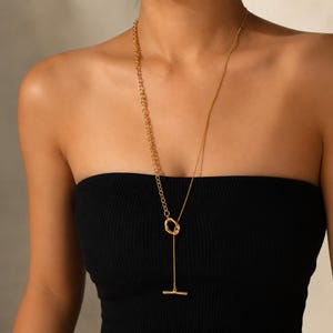 May include: A gold-toned lariat necklace featuring a long, thin chain on one side and a thicker, textured link chain on the other. The two chains meet at a small, irregular circular ring, with a single vertical chain ending in a horizontal bar.