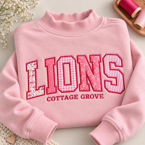 May include: A light pink long-sleeve sweatshirt with a mock neck collar. The front features the word LIONS in large, multi-patterned pink and white fabric letters, with the text COTTAGE GROVE embroidered in smaller pink letters below.