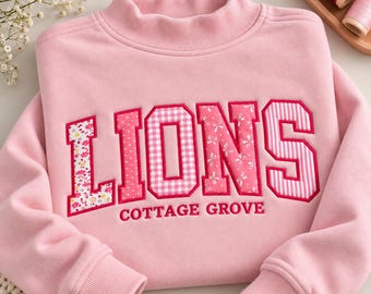 May include: A light pink long-sleeve sweatshirt with a mock neck collar. The front features the word LIONS in large, multi-patterned pink and white fabric letters, with the text COTTAGE GROVE embroidered in smaller pink letters below.