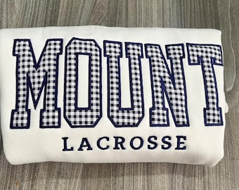 May include: A white sweatshirt featuring the word MOUNT in large, block-style letters filled with a navy blue and white gingham pattern. Below the word MOUNT, the word LACROSSE is embroidered in a smaller, solid navy blue sans-serif font.