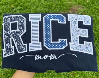 May include: A dark navy blue t-shirt featuring the word RICE in large block letters. Each letter has a unique pattern: floral, horizontal stripes, white polka dots on blue, and a light blue floral print. The word mom is written in white script below.