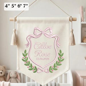 May include: A cream-colored fabric wall banner featuring an embroidered pink crest with a bow, green leaf accents, and the name Chloe Rose with the date 22.06.2026. The banner hangs from a wooden rod with tassels and text indicating 4" 5" 6" 7".