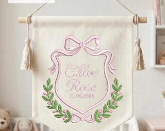 May include: A cream-colored fabric wall banner featuring an embroidered pink crest with a bow, green leaf accents, and the name Chloe Rose with the date 22.06.2026. The banner hangs from a wooden rod with tassels and text indicating 4" 5" 6" 7".