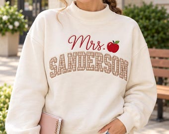 May include: An off-white long-sleeve sweatshirt featuring the embroidered text Mrs. Sanderson in red script and brown plaid block letters, accompanied by a small red apple graphic with a green leaf on the upper chest area.