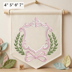 May include: A cream-colored fabric wall banner featuring an embroidered pink decorative frame with a central shield shape, flanked by green laurel branches and a pink bow at the base. The banner includes text indicating sizes 4", 5", 6", 7".