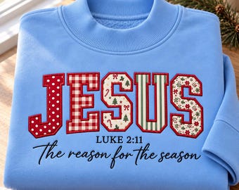 May include: A light blue crewneck sweatshirt featuring the word JESUS in large, patterned block letters with red outlines. Below, the text reads LUKE 2:11 and The reason for the season in black script. The letters feature polka dots, plaid, and holiday motifs.