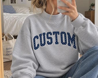 May include: A light gray crewneck sweatshirt featuring the word CUSTOM in dark blue block letters with white polka dots. The sweatshirt has long sleeves, a ribbed collar, and a relaxed fit, worn with blue denim jeans.