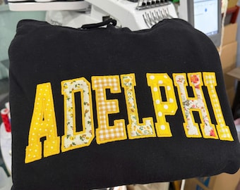 May include: A black hooded sweatshirt featuring the word ADELPHI in large, block-style letters. Each letter is outlined in black and filled with a variety of yellow patterns, including polka dots, gingham checks, and floral prints.