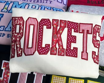 May include: A white sweatshirt featuring the word ROCKETS in large, collegiate-style block letters. Each letter is appliqued with a different red-toned fabric pattern, including floral prints, polka dots, and vertical stripes, all outlined in red.