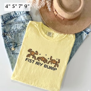 May include: A pale yellow t-shirt featuring a graphic of three brown crabs with the text FIST MY BUMP, displayed alongside acid-wash blue jeans, a tan wide-brimmed hat, and a text overlay indicating sizes 4, 5, 7, and 9 inches.