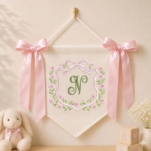 May include: A cream-colored fabric wall banner featuring an embroidered green letter N centered within a pink floral wreath and ribbon design. The banner is suspended by a white cord and adorned with two large, light pink satin bows on each side.