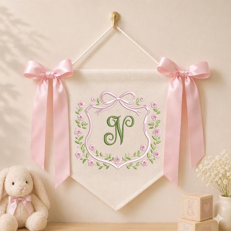 May include: A cream-colored fabric wall banner featuring an embroidered green letter N centered within a pink floral wreath and ribbon design. The banner is suspended by a white cord and adorned with two large, light pink satin bows on each side.