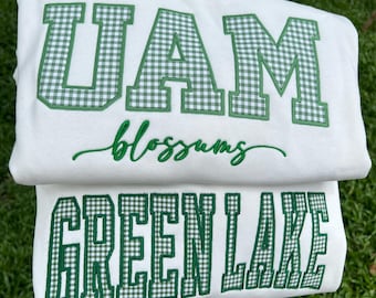May include: A white sweatshirt featuring the letters UAM in a green and white gingham pattern with a thick green border. Below the letters, the word blossoms is embroidered in green cursive script. Below that, the words GREEN LAKE appear.