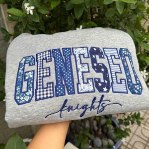 May include: A heather gray sweatshirt featuring the word GENESEO in large, block-style letters. Each letter is filled with a different blue and white patterned fabric, including floral, gingham, polka dot, and star prints. The word knights is embroidered below in blue cursive script.