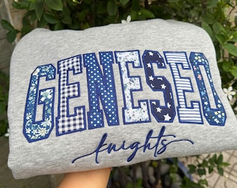 May include: A light gray sweatshirt featuring the word GENESEO in large, block-style letters. Each letter is filled with a different blue and white patterned fabric, including floral, gingham, polka dot, and star prints. The word knights is embroidered below in blue script.