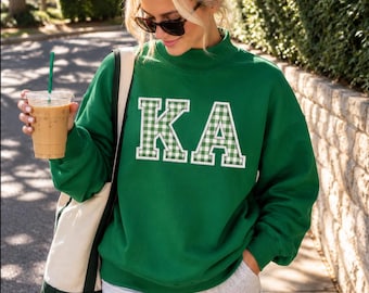 May include: A forest green long-sleeve sweatshirt featuring a mock neck and the letters KA in white and green gingham check applique. The sweatshirt is paired with light gray shorts and a cream and green canvas tote bag.