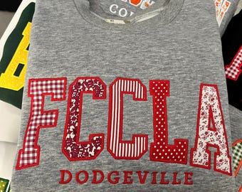 May include: A heather gray crewneck sweatshirt featuring the letters FCCLA in large, red-bordered applique patches with varied patterns including red gingham, floral, striped, and polka dot prints, with the word DODGEVILLE embroidered in red below.