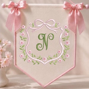 May include: A cream-colored fabric wall banner featuring a central embroidered green letter N. The banner is framed by a pink embroidered crest and floral wreath, finished with pink satin ribbon bows at the top corners on a wooden rod.